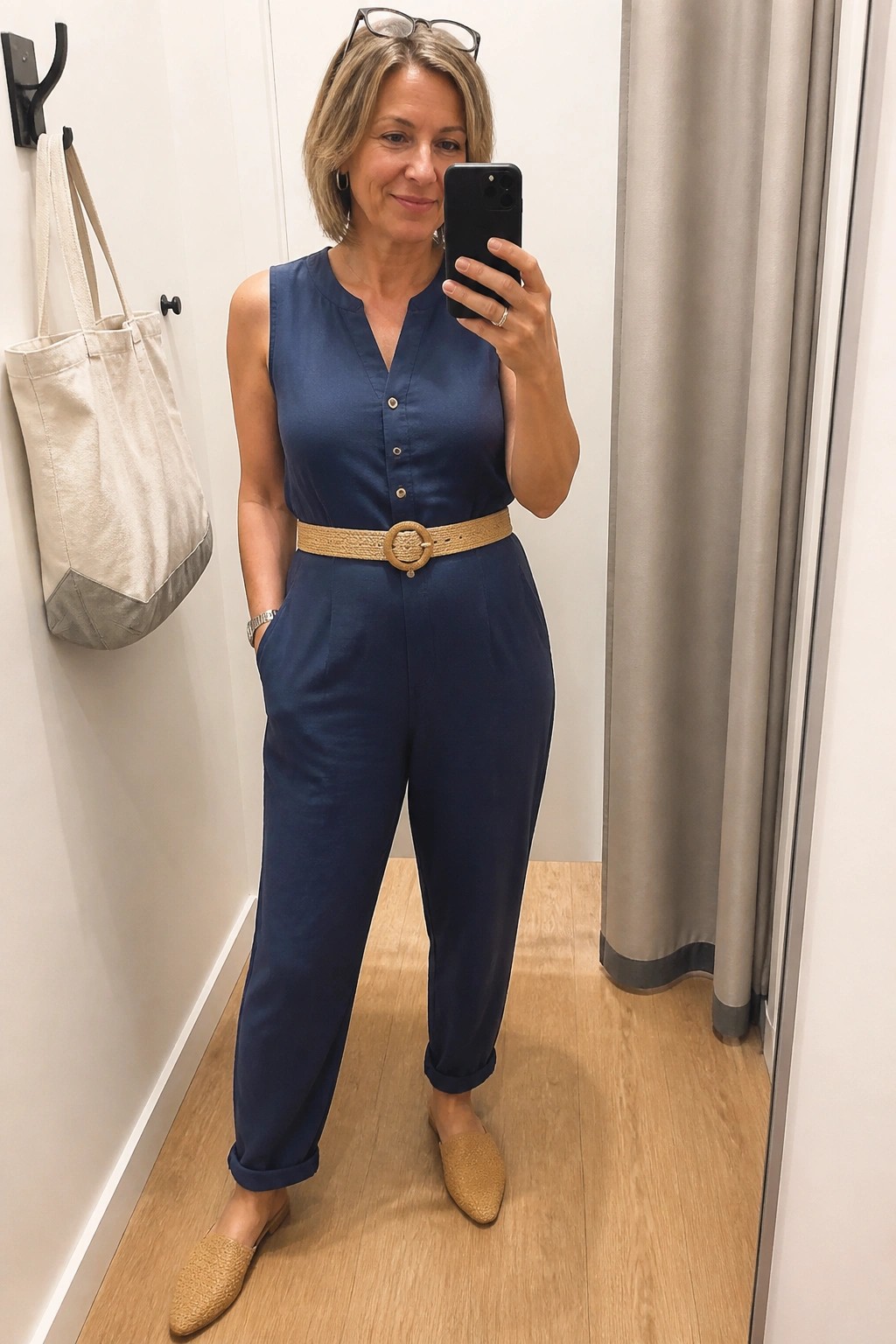 Woman in navy jumpsuit with woven belt taking mirror selfie in fitting room.