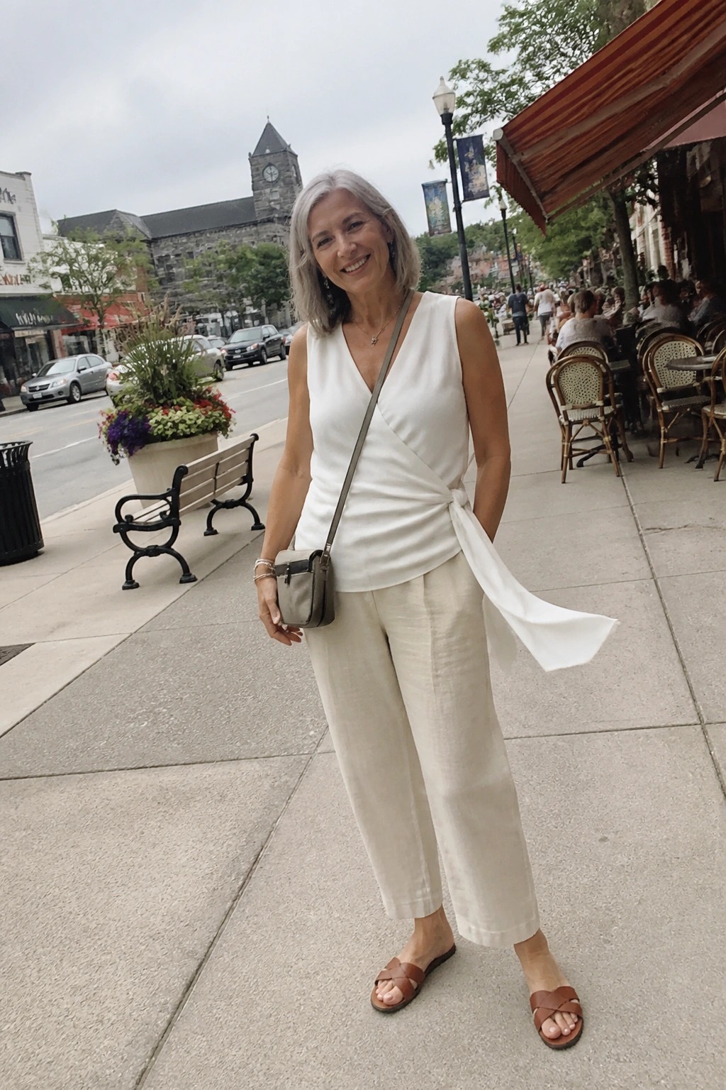 Smiling gray-haired woman in white wrap top and beige pants on cafe-lined sidewalk