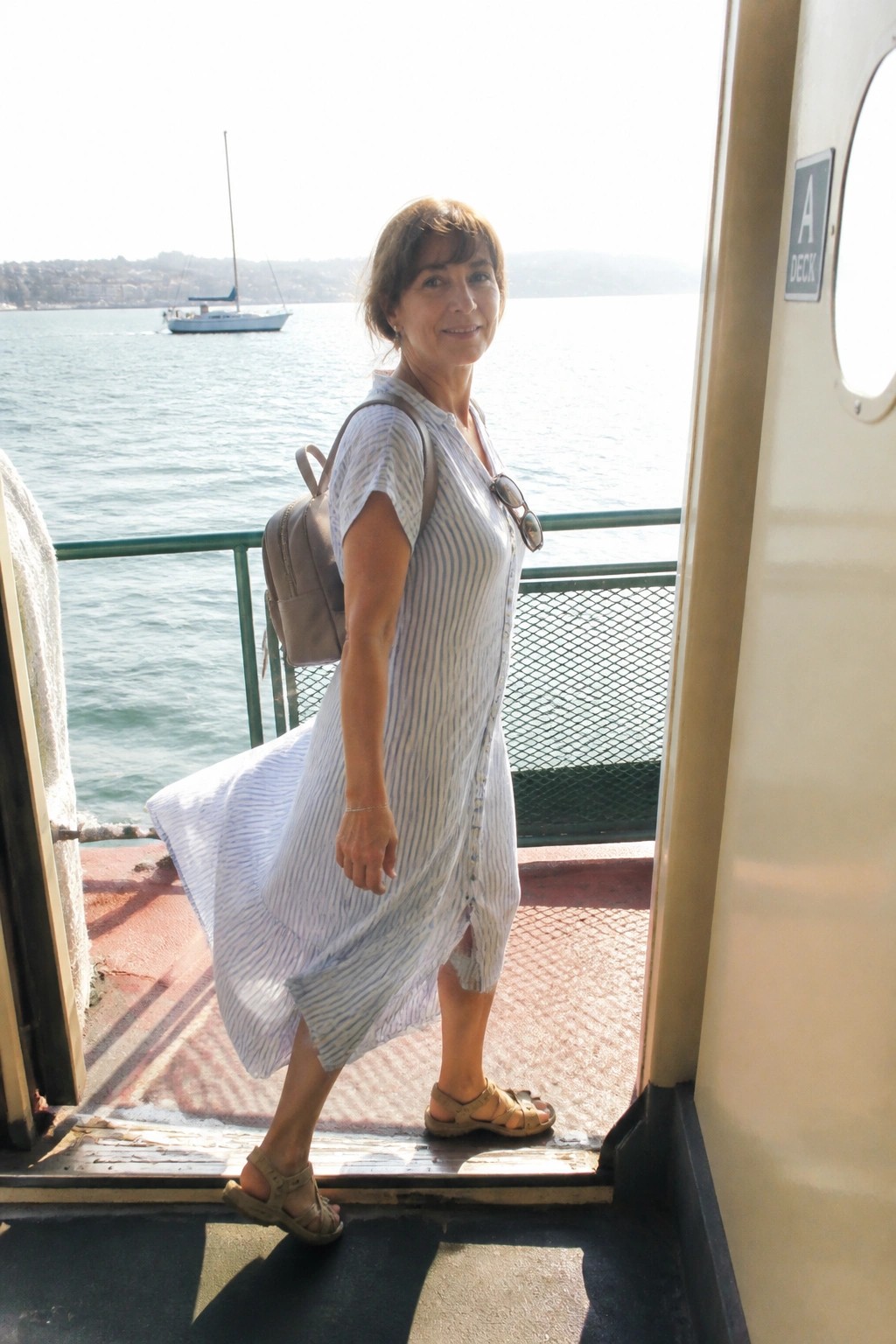 Woman in blue striped dress with backpack on boat deck near sailboat