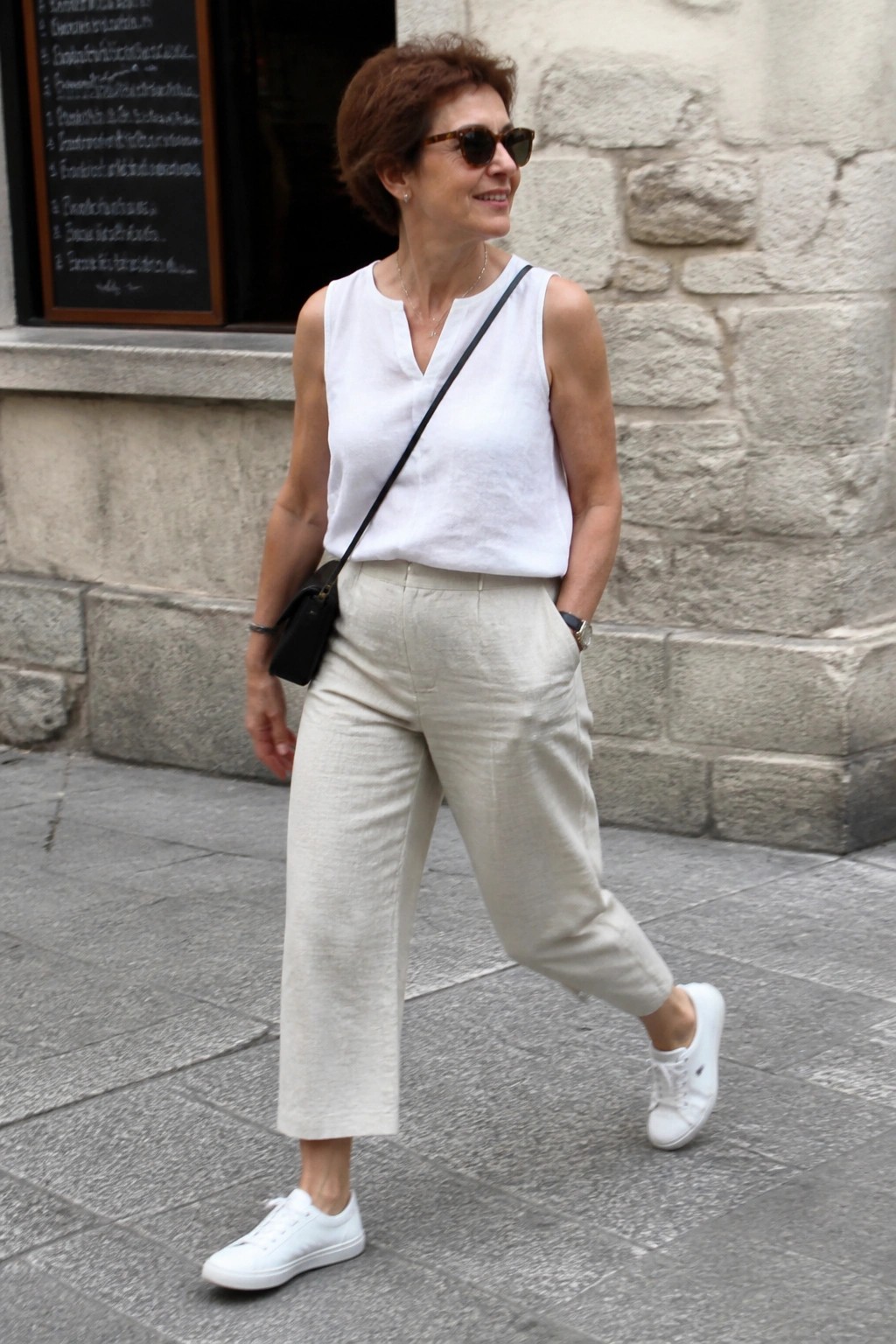 Woman in white top and beige pants with black bag walking on stone street