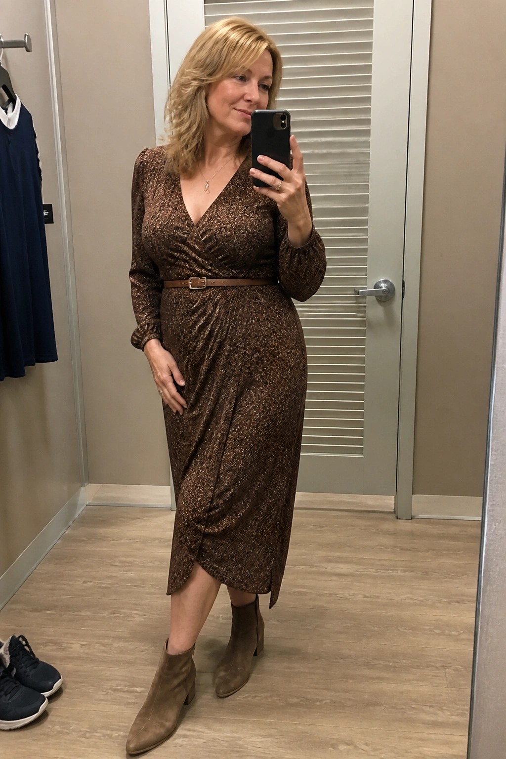 Woman in brown sparkly wrap dress and boots taking mirror selfie in fitting room