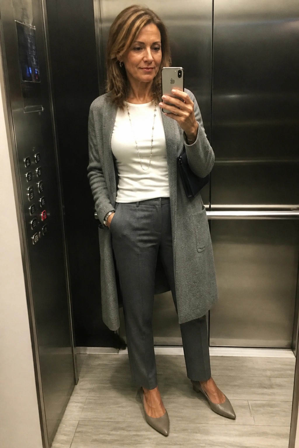 Woman in gray cardigan and trousers taking selfie in elevator.
