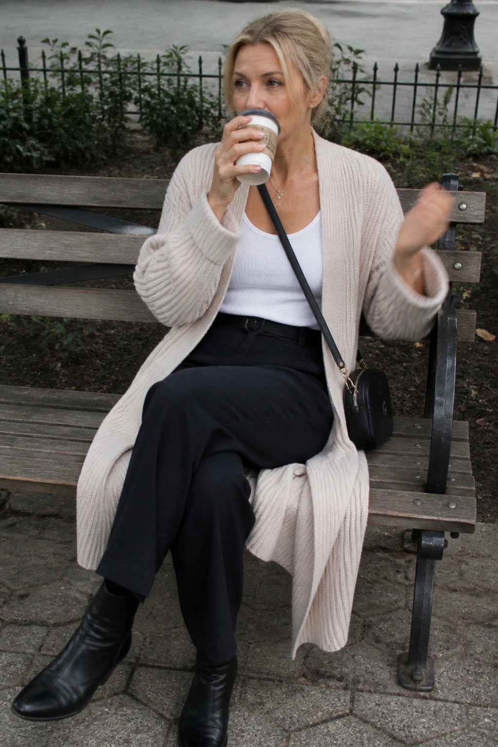 Blonde woman in beige cardigan sips coffee on wooden bench, gesturing with hand.