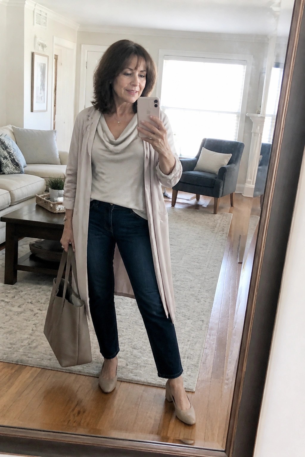 Woman in beige cardigan and dark jeans taking mirror selfie with tote bag.