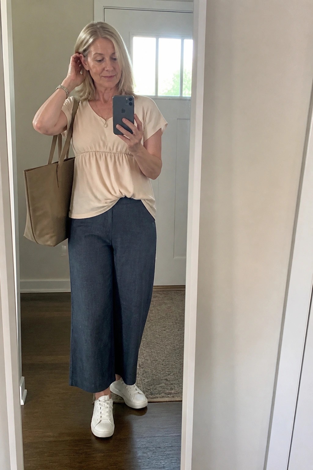 Blonde woman in beige peplum top and blue wide-leg pants holding phone and brown tote bag