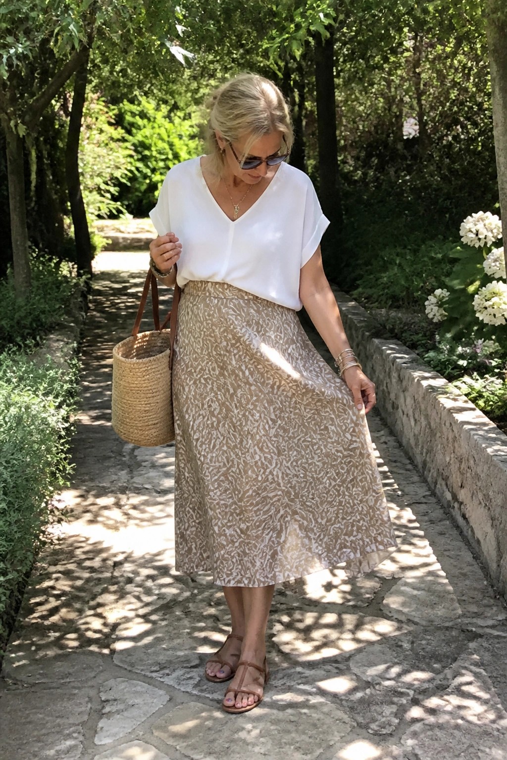 Woman in white top and patterned beige skirt holding woven bag on garden path