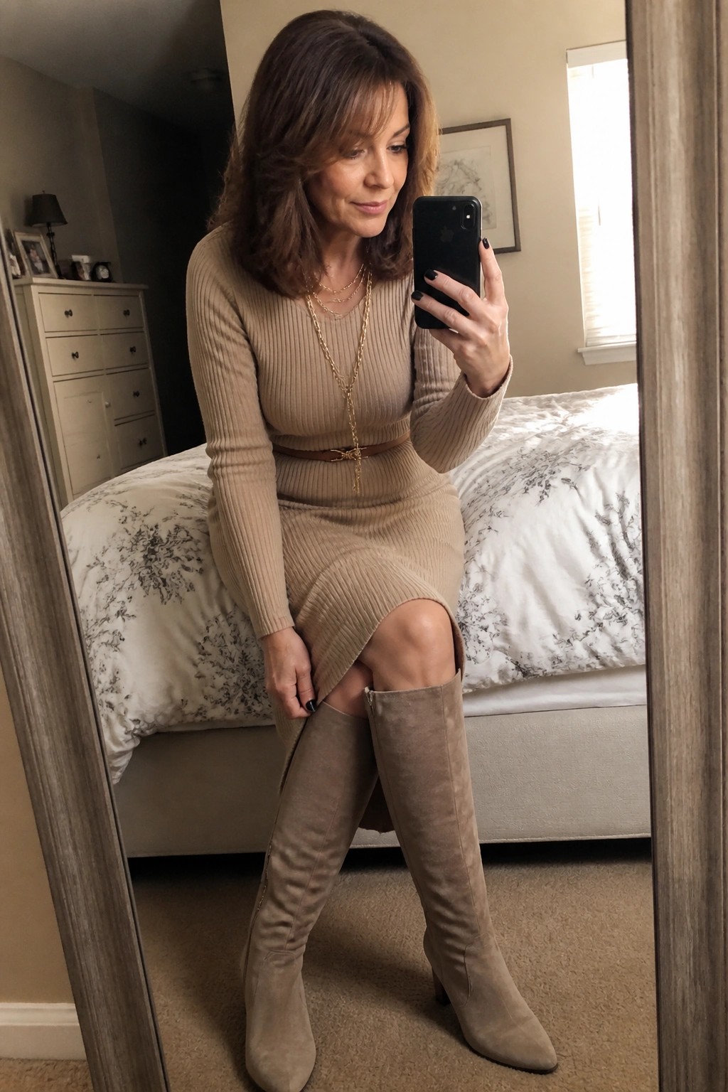 Woman in beige ribbed dress and suede boots taking bedroom mirror selfie
