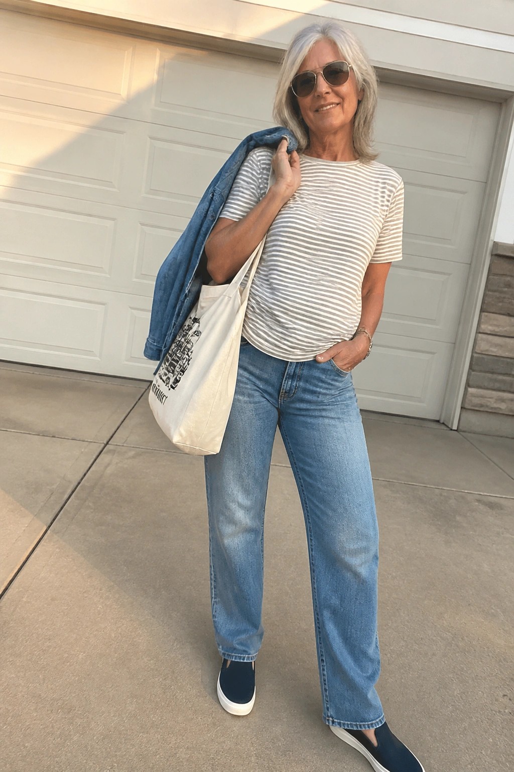 Woman over 50 in striped short sleeve tee tucked into blue wide leg jeans with denim jacket over one shoulder, white tote bag, navy slip on shoes and sunglasses standing in front of garage door.