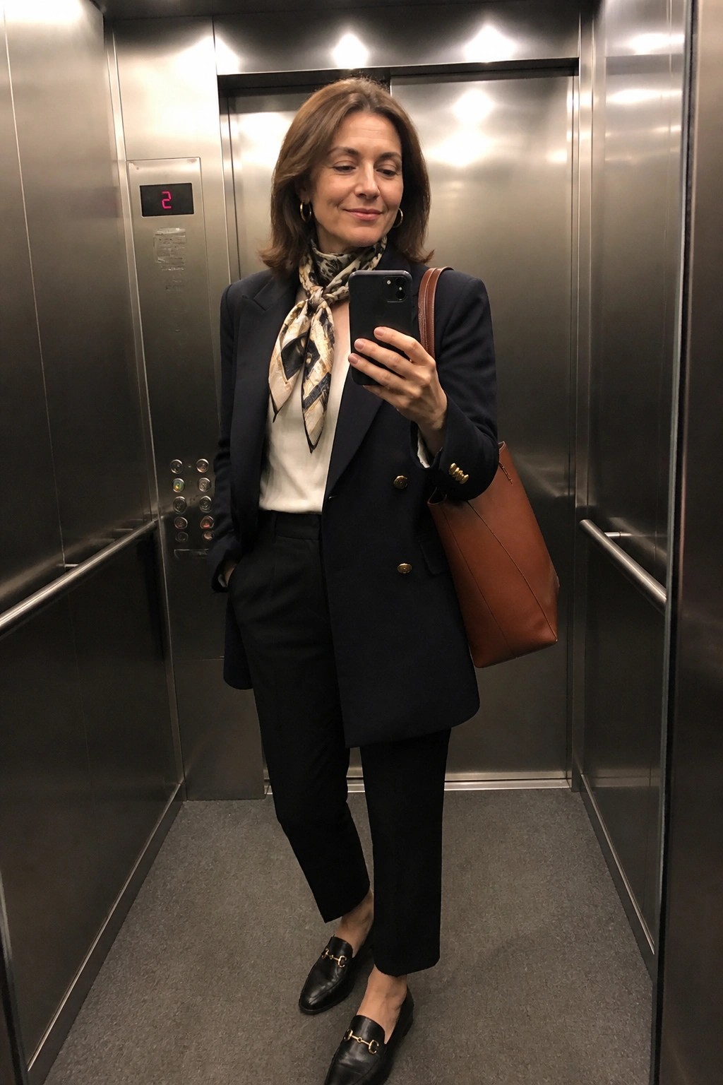 Woman in navy blazer and patterned scarf takes selfie in elevator.