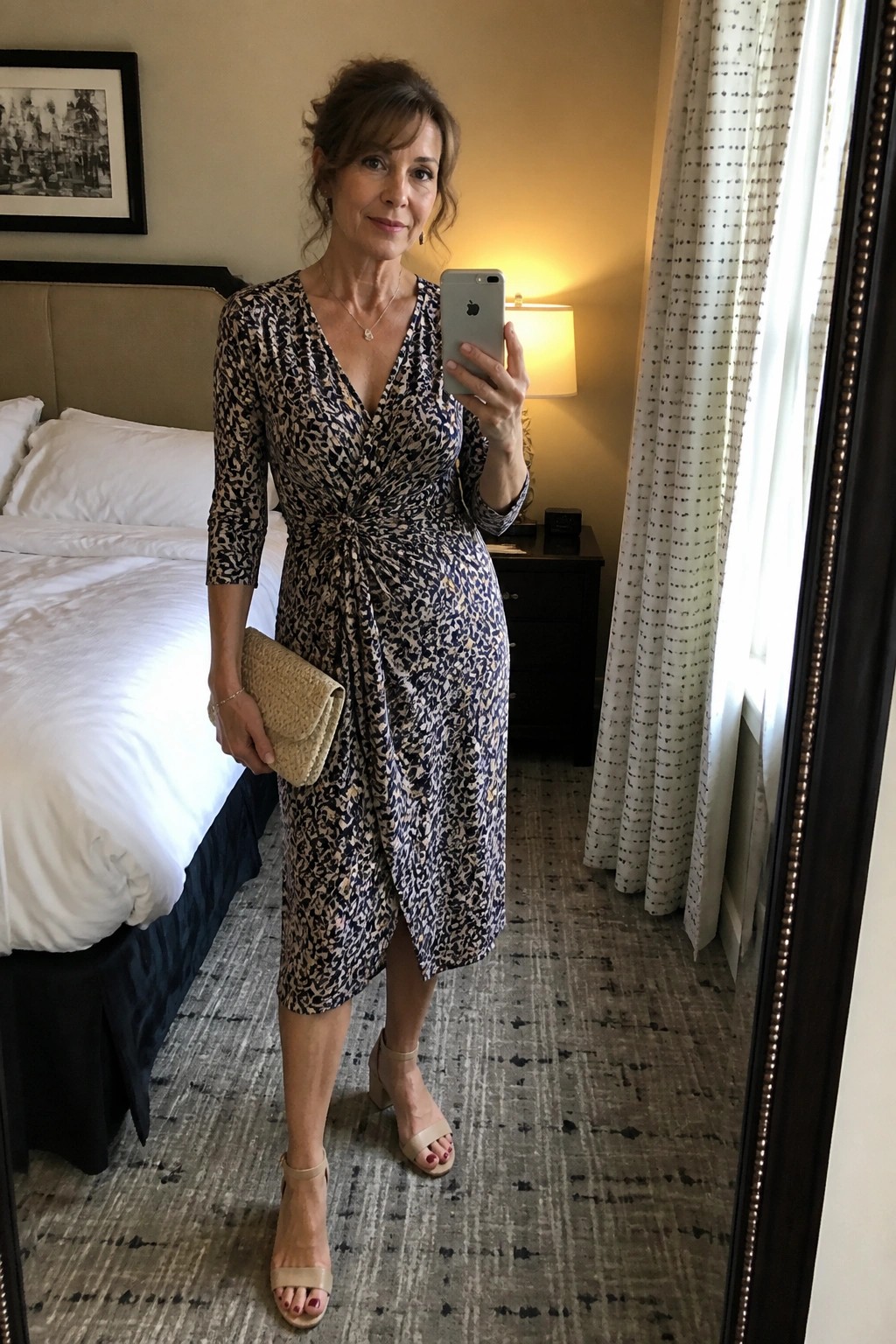 Woman in patterned wrap dress taking mirror selfie with clutch in hotel room
