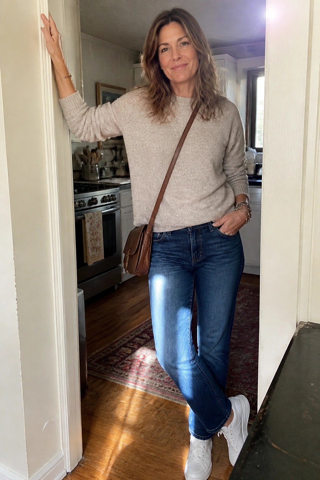 Woman in beige sweater and jeans leaning in kitchen doorway with brown bag.