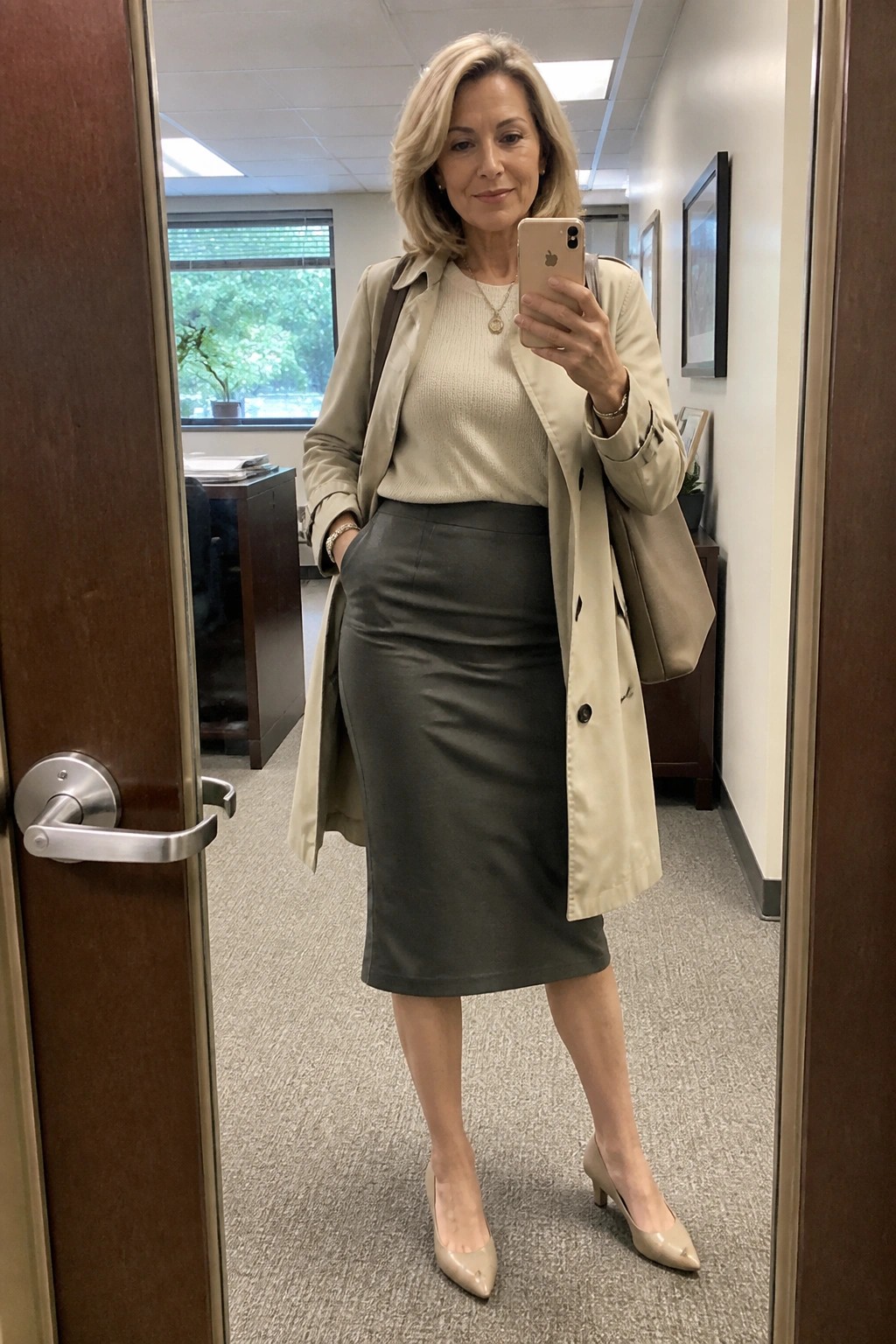 Woman in beige trench coat and gray skirt taking mirror selfie in office hallway