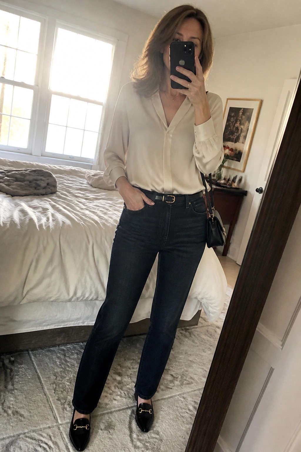 Woman in cream blouse and dark jeans taking mirror selfie in bedroom