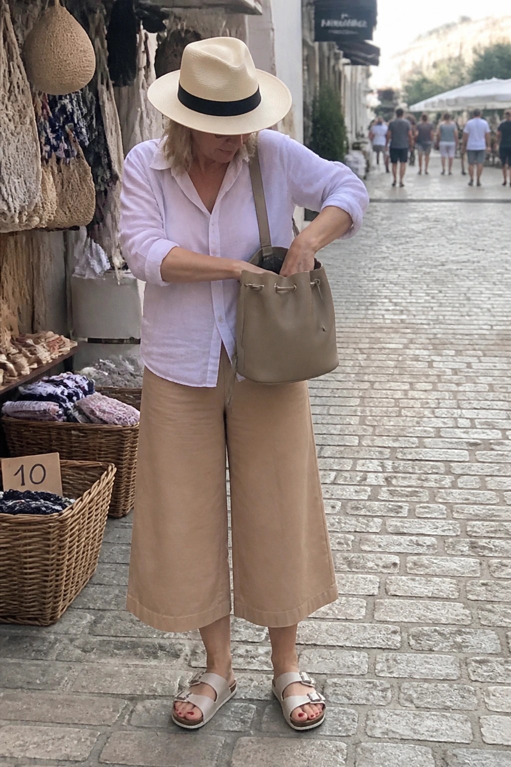 Woman in pink shirt and beige culottes reaching into taupe bucket bag on cobblestone street
