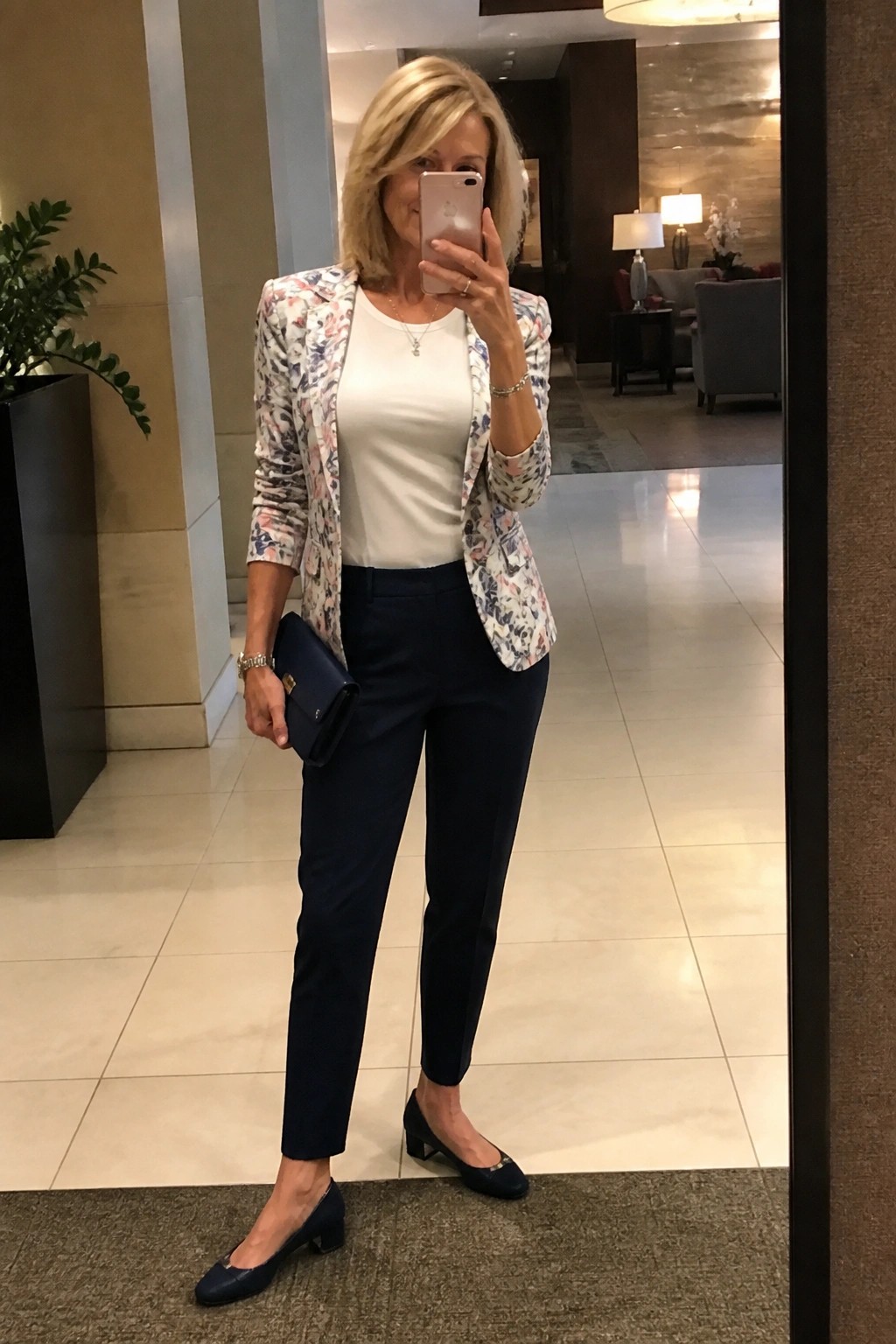 Woman wearing a white floral print blazer over a white top with navy trousers navy flats and a navy clutch in a modern lobby setting.