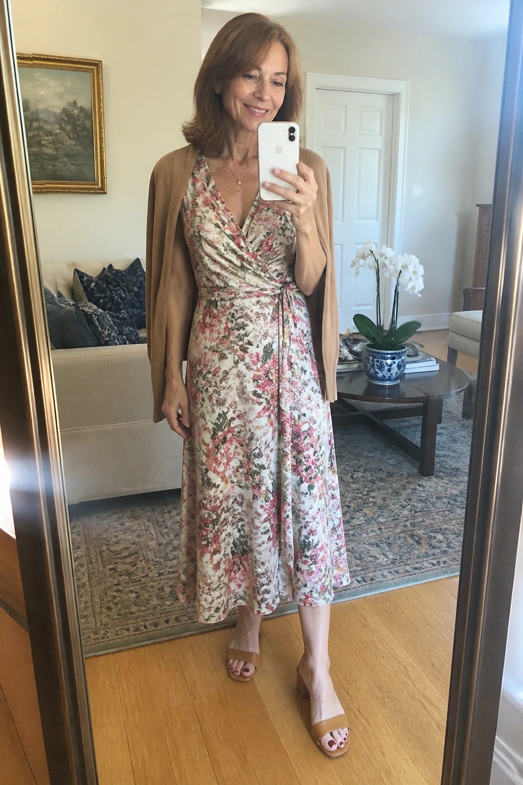 Woman in floral wrap dress and tan cardigan taking mirror selfie in living room