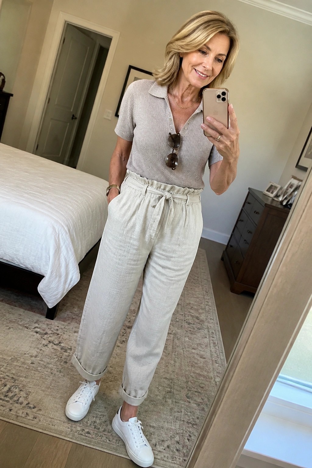 Blonde woman in beige polo and cuffed linen pants taking bedroom mirror selfie