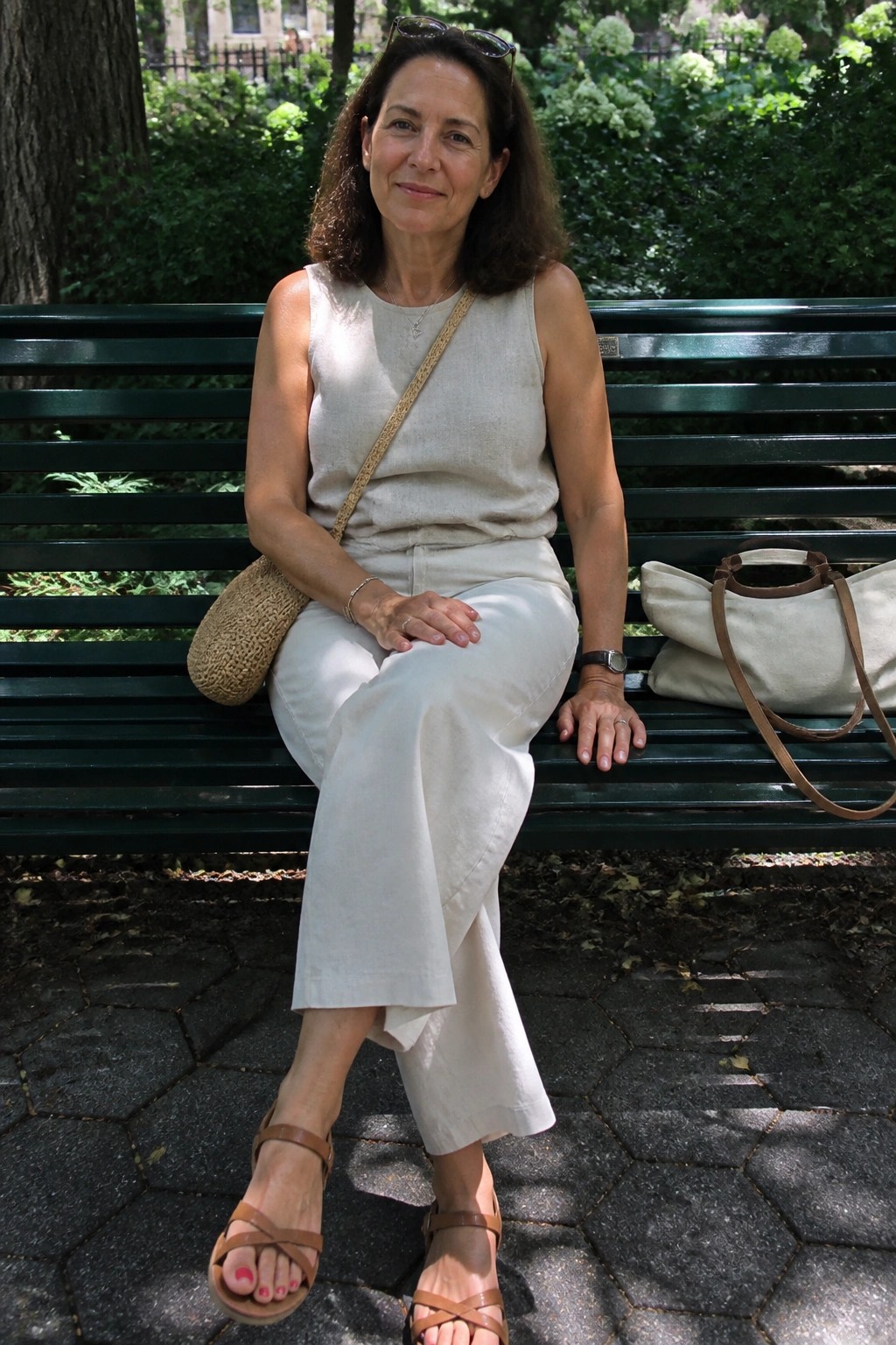 Woman in beige outfit seated on green park bench with bags nearby