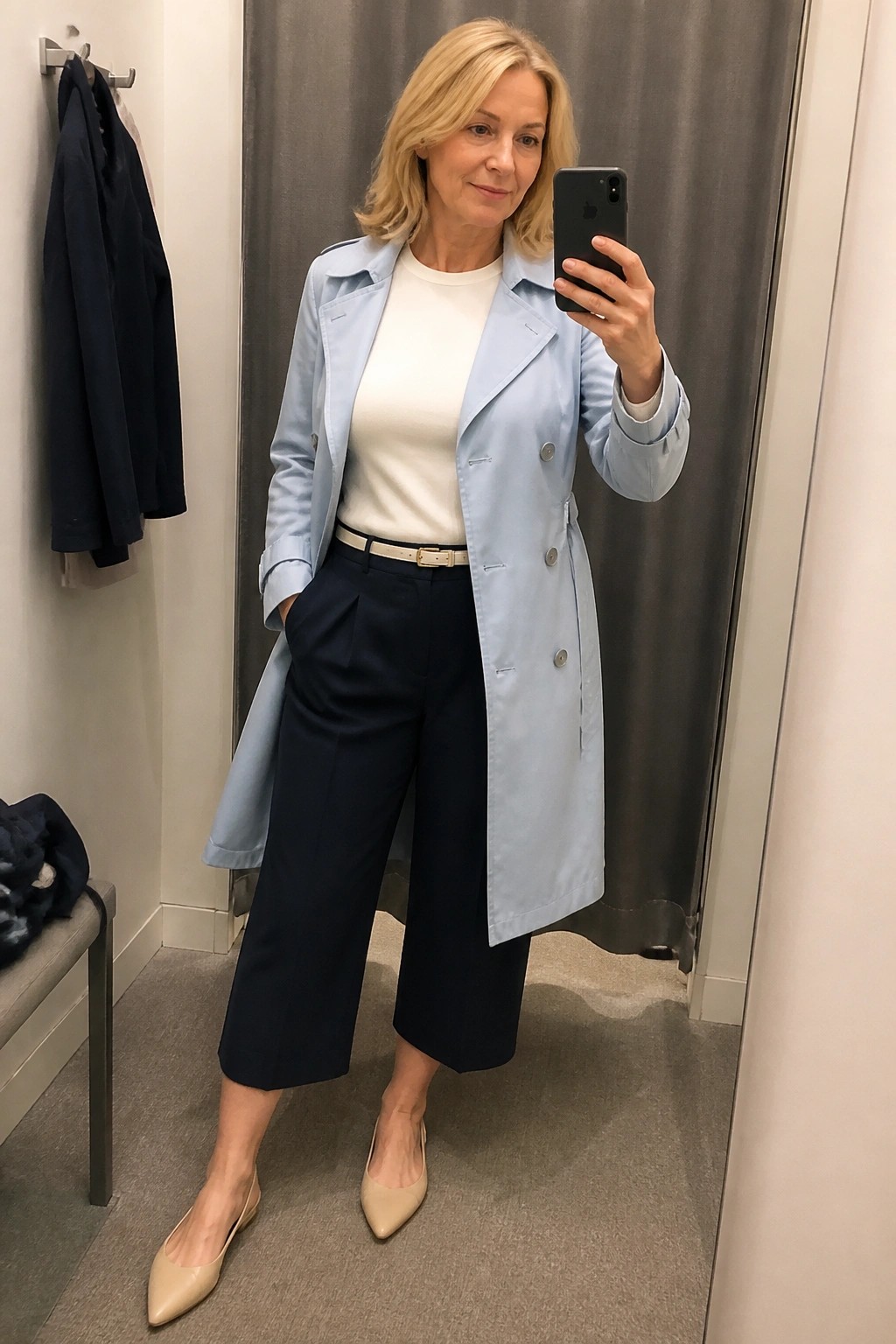 Woman over 50 wearing a light blue trench coat open over a white top with a white belt navy wide leg cropped pants and beige pointed flats taking a mirror selfie in a fitting room.