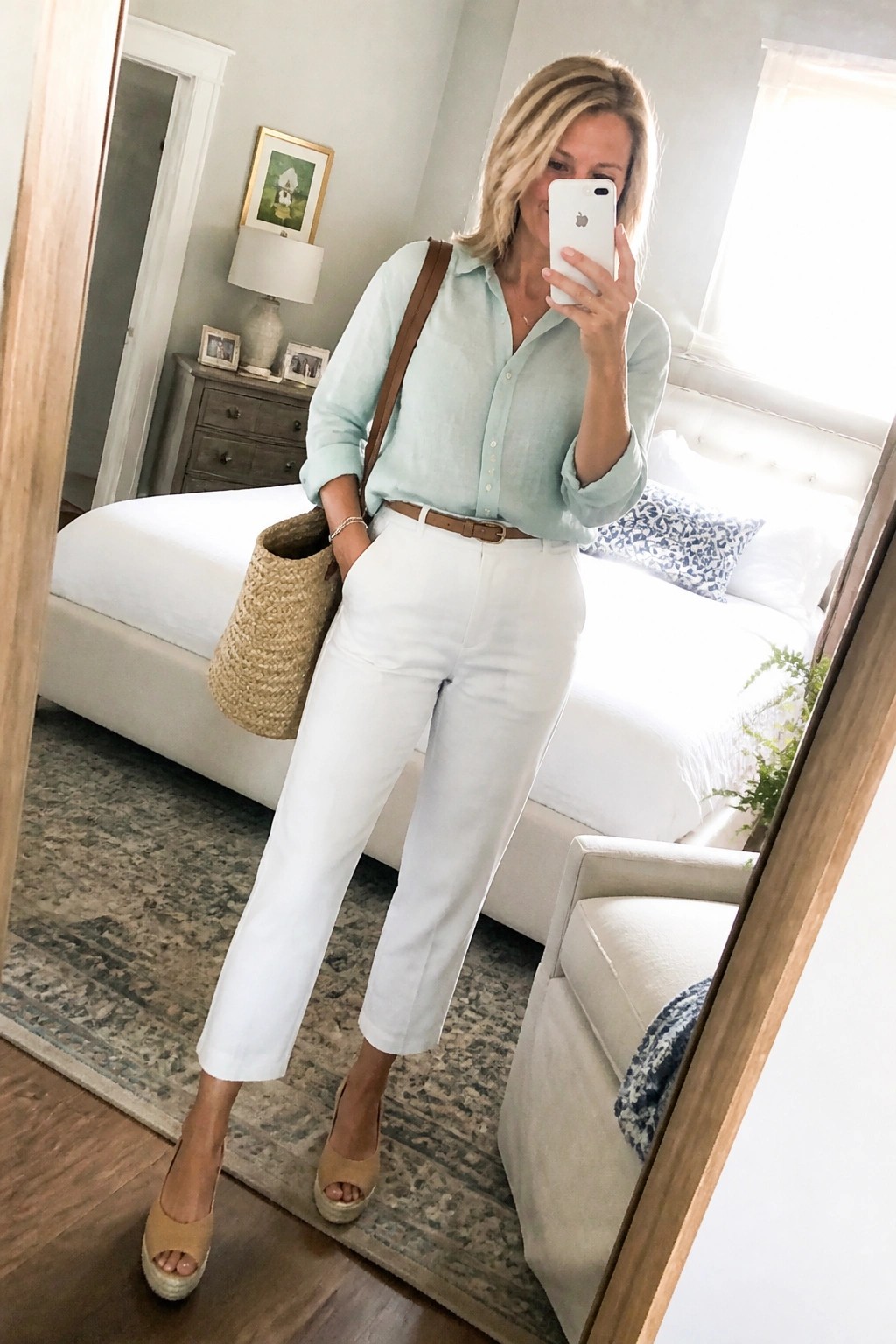 Woman in light blue shirt and white pants taking mirror selfie in bedroom