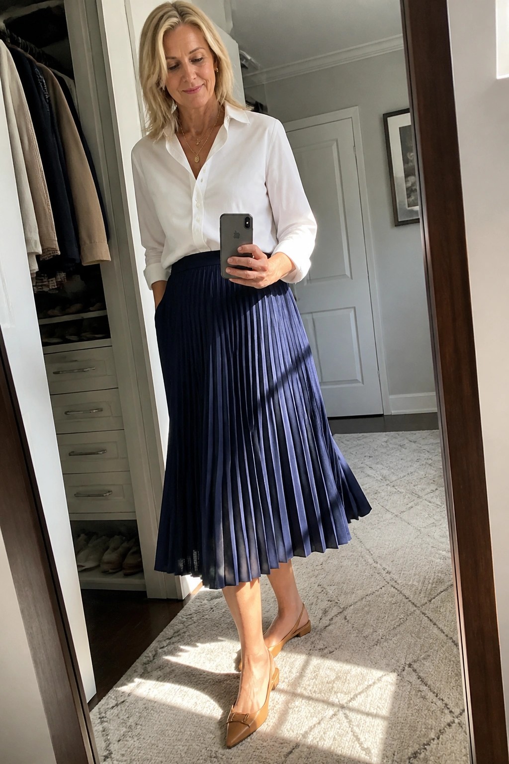 Woman over 50 wearing a white button down shirt tucked into a navy pleated midi skirt with brown pointed flats and layered gold necklaces while holding a phone in a mirror selfie.