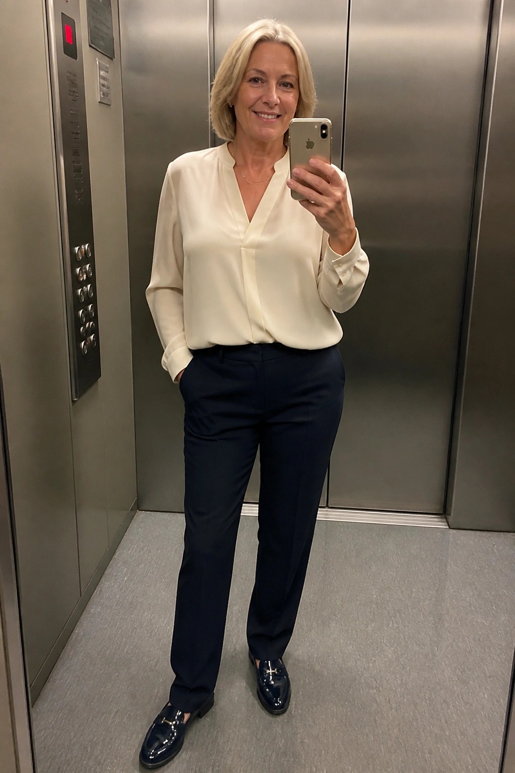 Smiling blonde woman in cream blouse and navy pants taking selfie in elevator