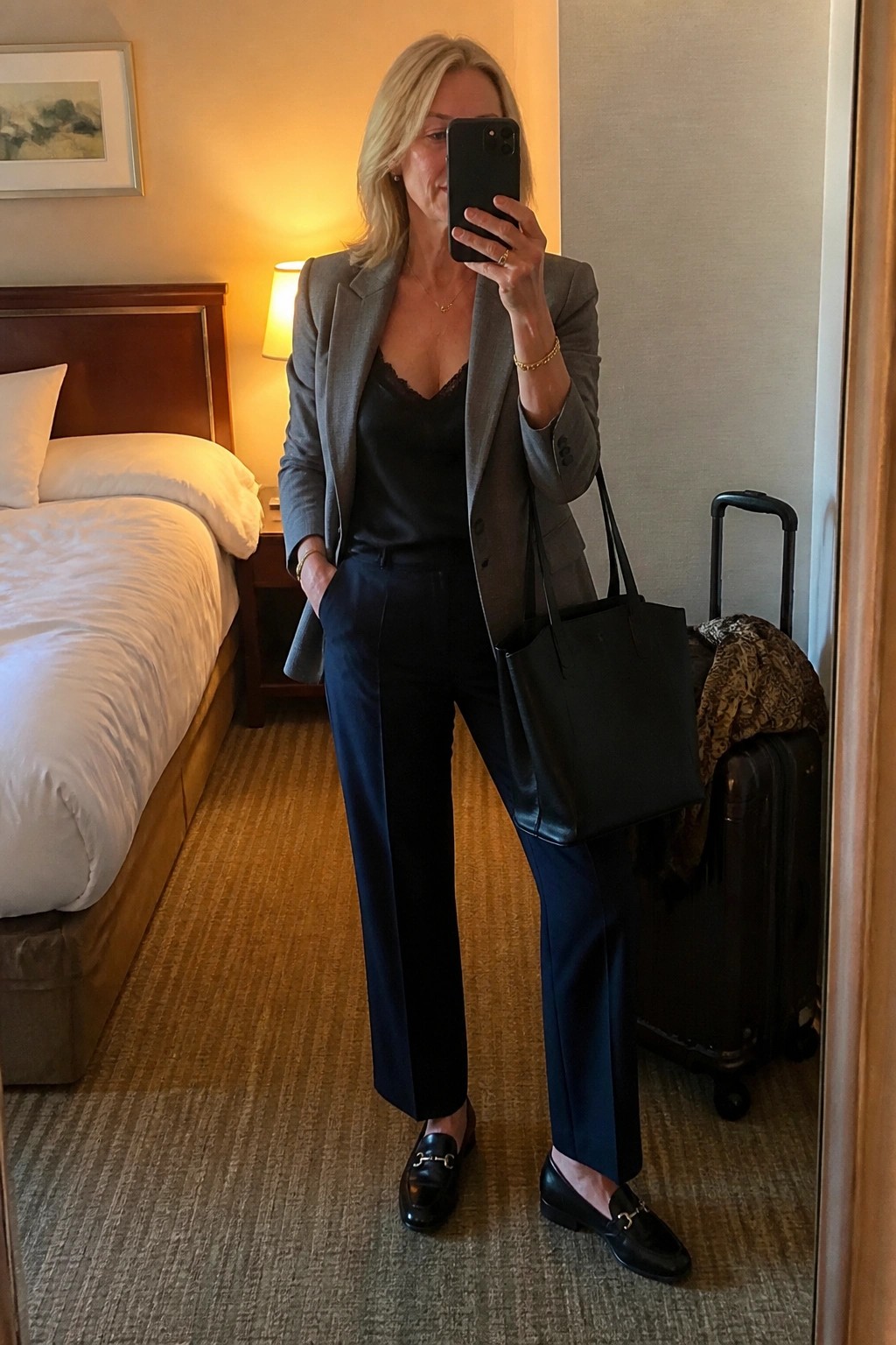 Blonde woman in gray blazer and navy pants taking mirror selfie in hotel room