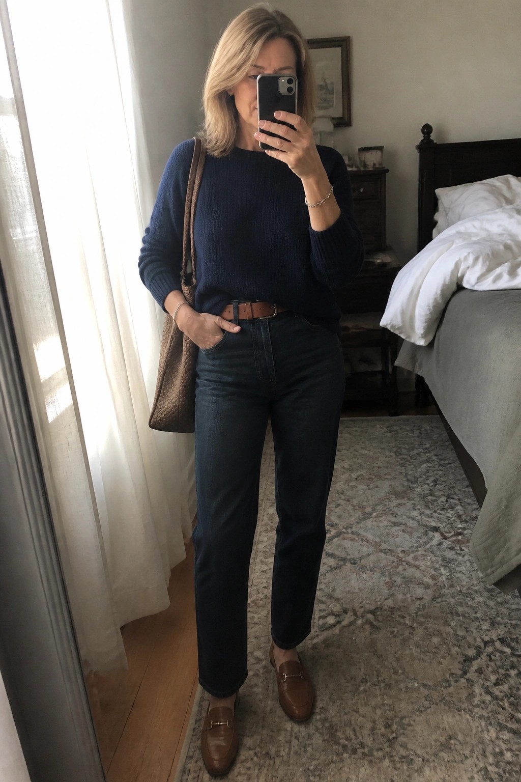 Woman over 50 wearing a navy ribbed sweater tucked into dark straight leg jeans with a brown leather belt, brown loafers, and a brown woven tote bag, standing in a bedroom.