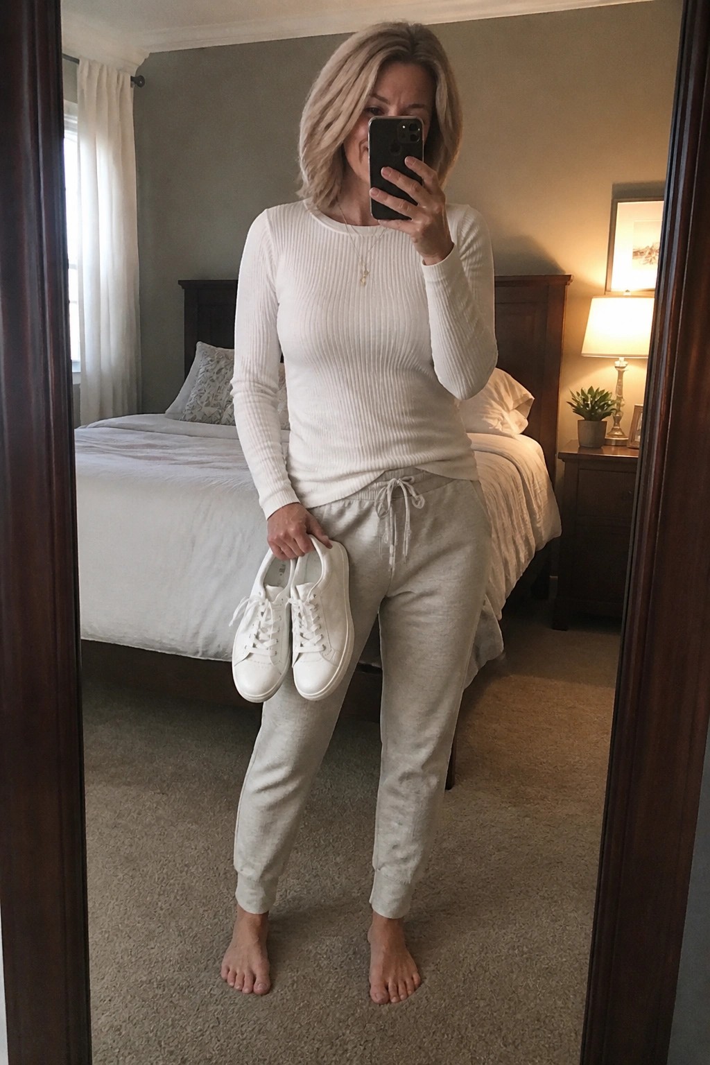 Woman in white top and gray sweatpants holding sneakers for mirror selfie in bedroom