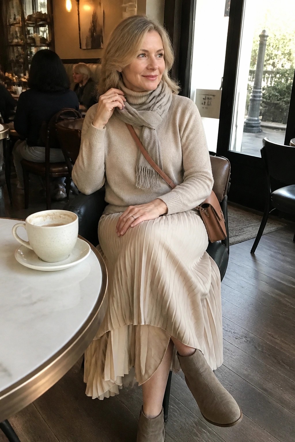 Elegant blonde woman in beige turtleneck and pleated skirt at cafe table with coffee