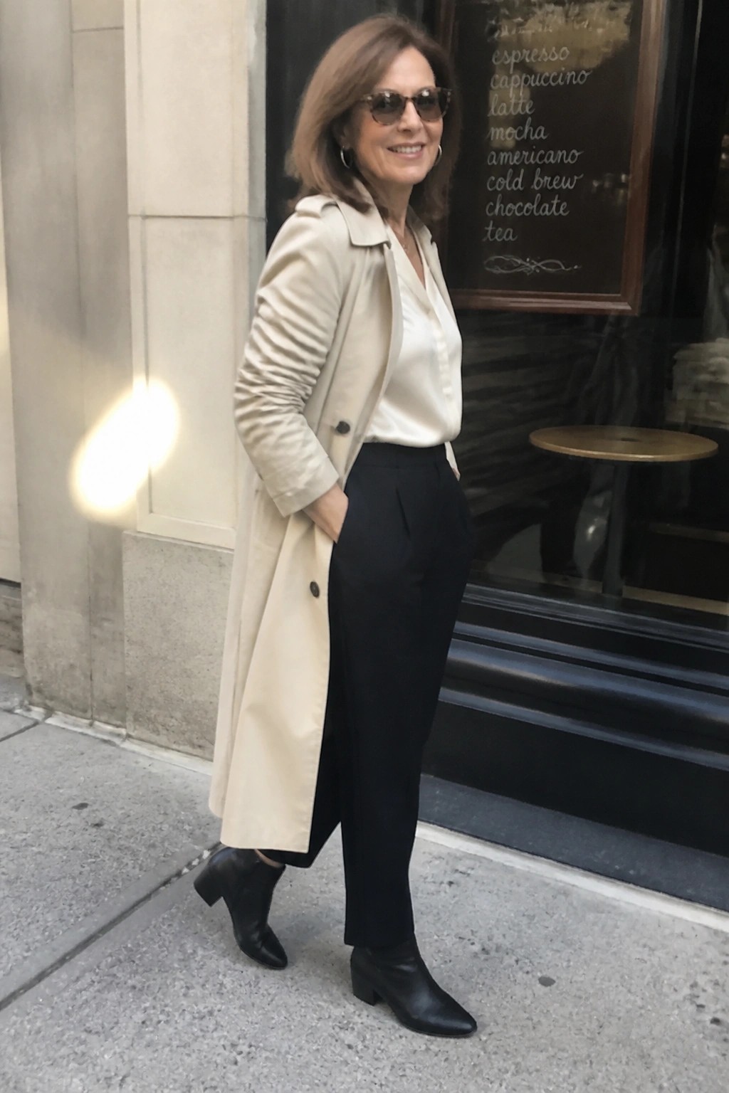 Woman in beige trench coat, white blouse, black pants, and boots outside cafe