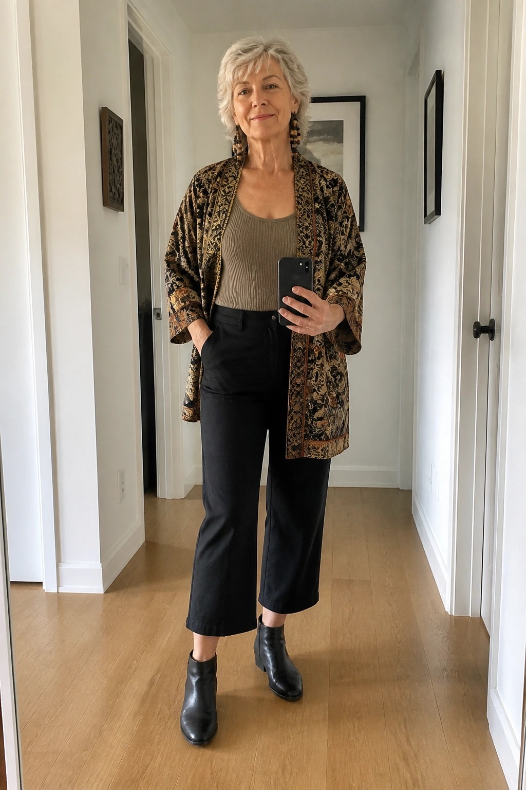 Woman with short gray hair in patterned jacket, beige top, and black pants holding phone.