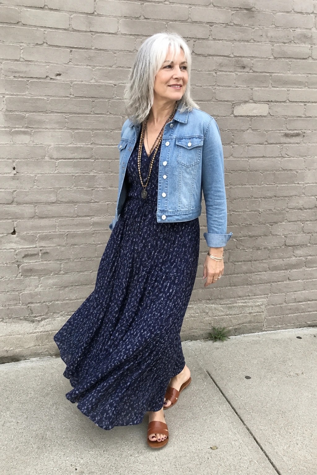 Woman wearing light blue denim jacket over dark blue patterned maxi dress with layered necklaces and brown flat sandals.