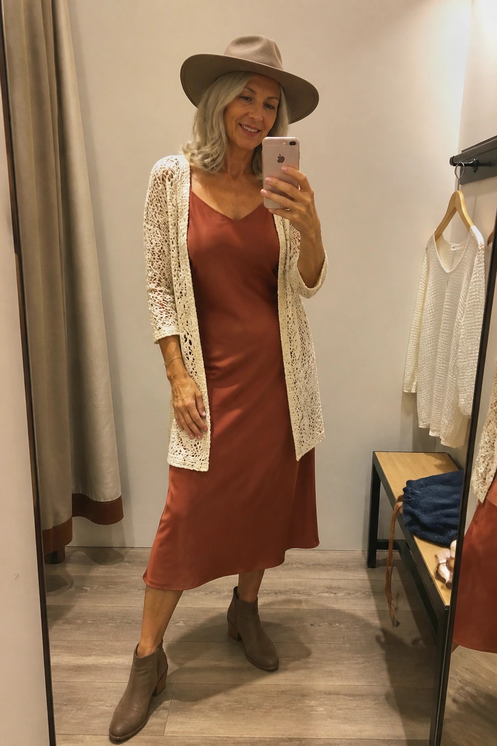 Woman in rust slip dress, white crochet cardigan, and hat taking mirror selfie