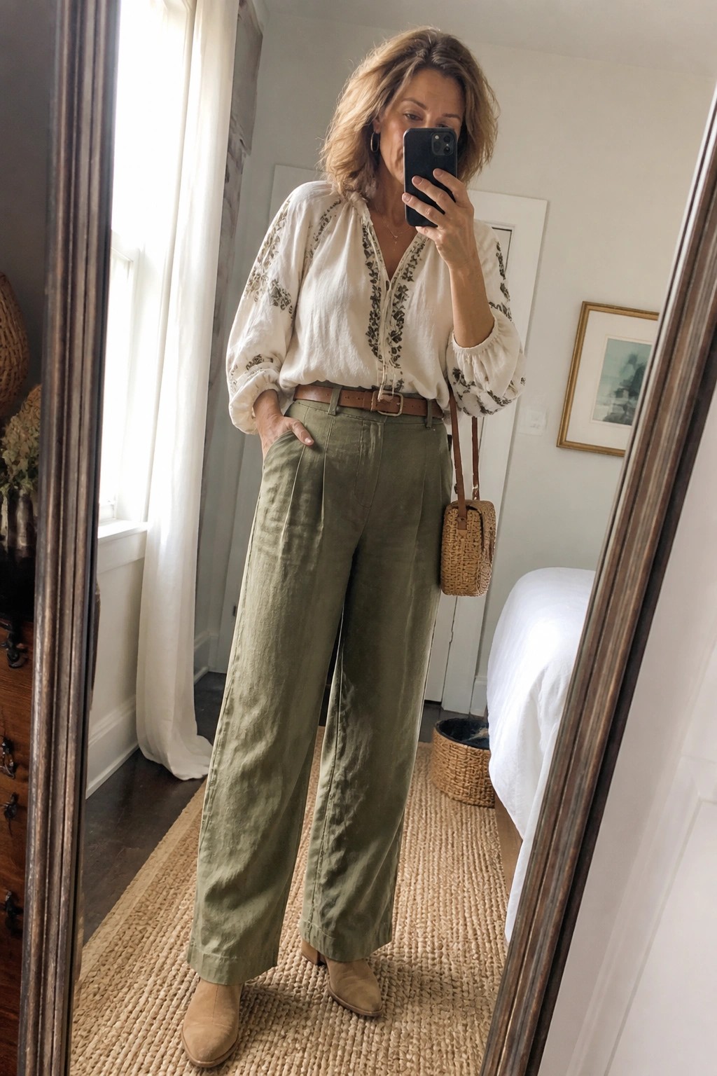 Woman in white embroidered blouse and green wide-leg pants taking bedroom mirror selfie