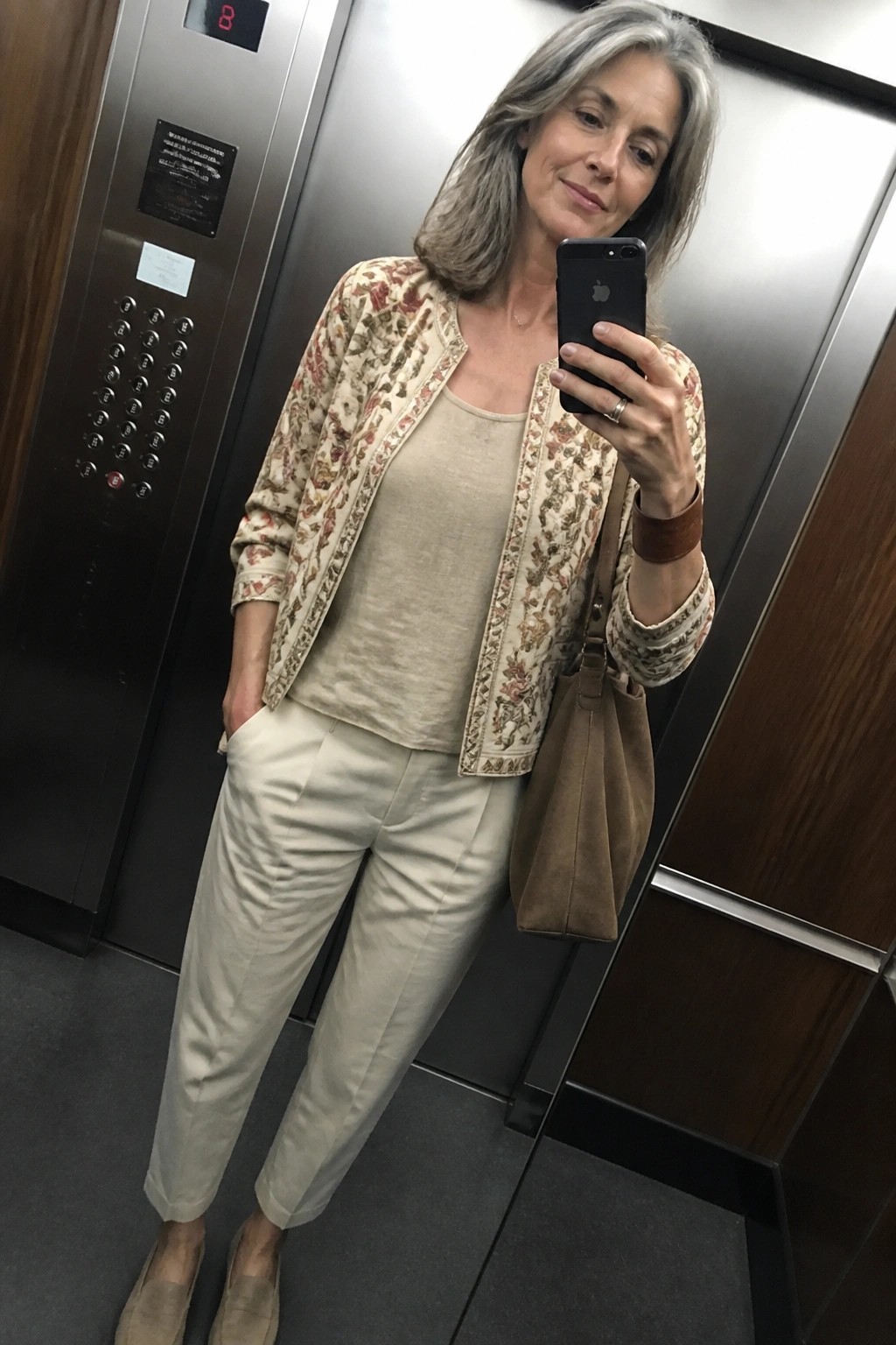 Woman taking a mirror selfie in an elevator wearing a cream floral embroidered jacket over a beige linen tank and matching straight pants, paired with suede loafers and a brown suede shoulder bag.