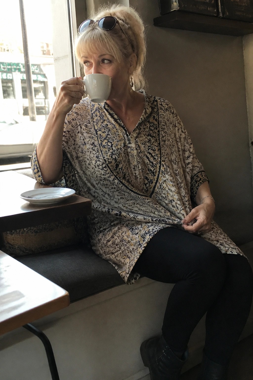 Blonde woman in patterned tunic sips coffee by cafe window
