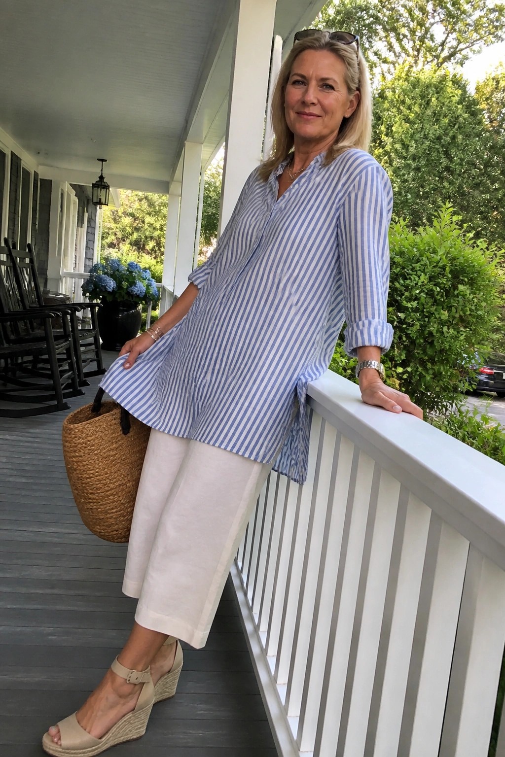 Blonde woman in blue striped tunic and white pants on porch with straw bag.