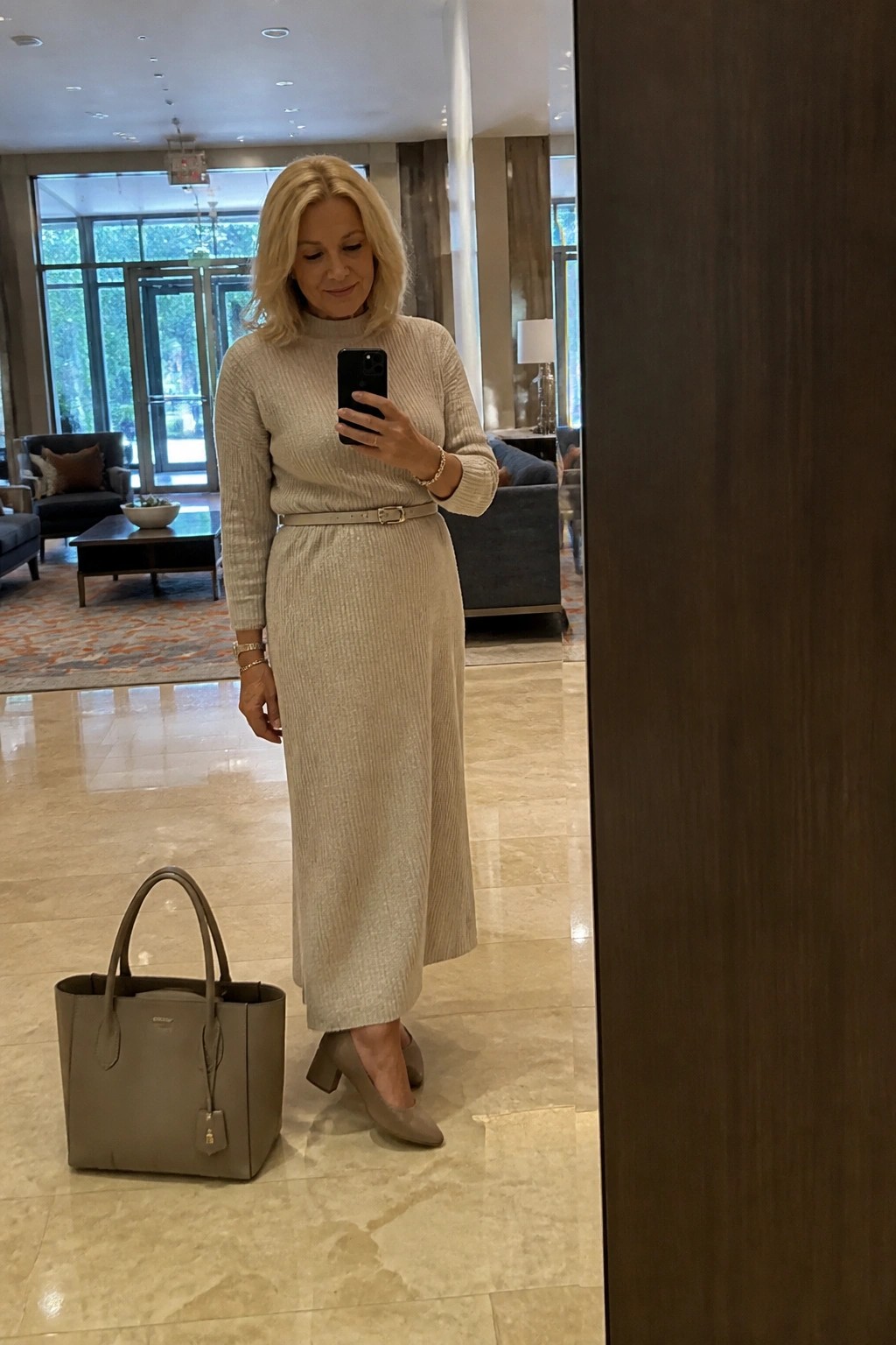 Blonde woman in beige ribbed knit dress taking mirror selfie beside taupe handbag