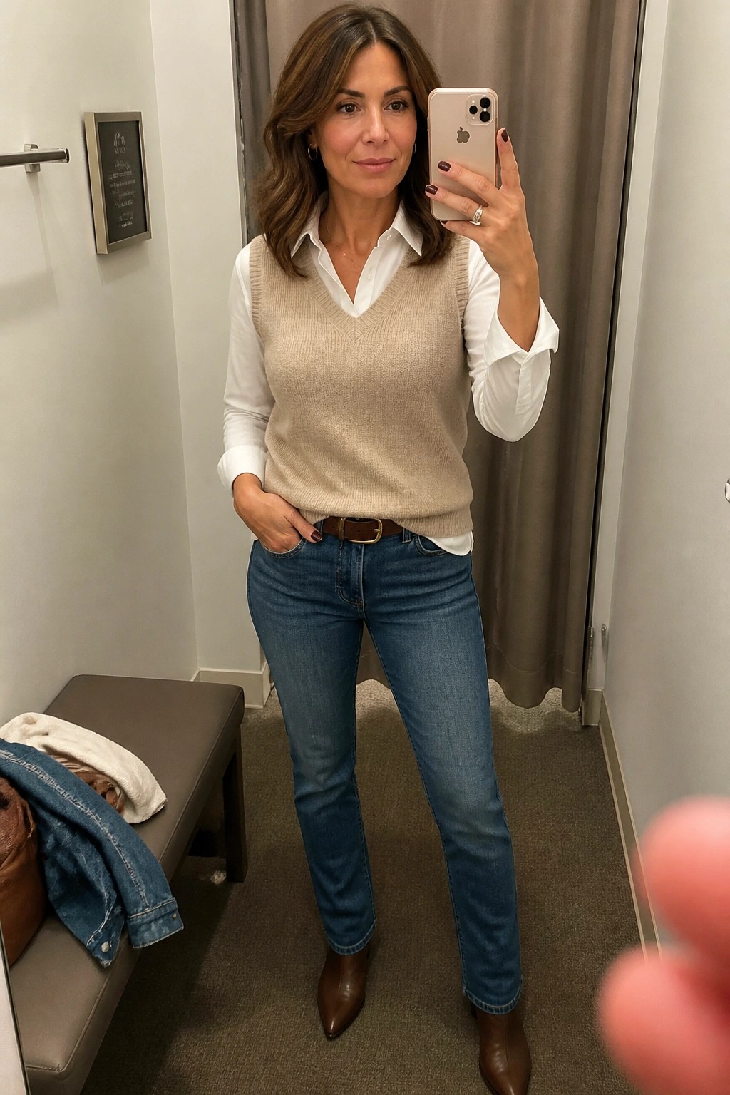 Woman in beige sweater vest, white shirt, and blue jeans taking mirror selfie in fitting room.