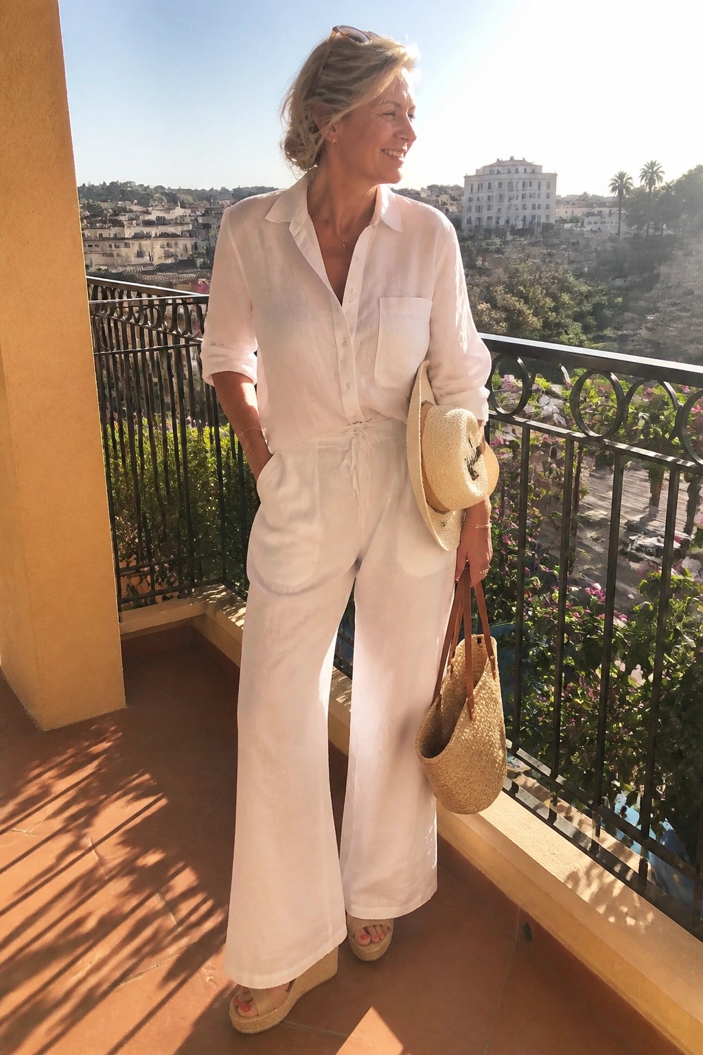 Woman in white linen button front shirt tied at waist with matching wide leg pants, holding straw hat and woven tote while wearing espadrille wedge sandals