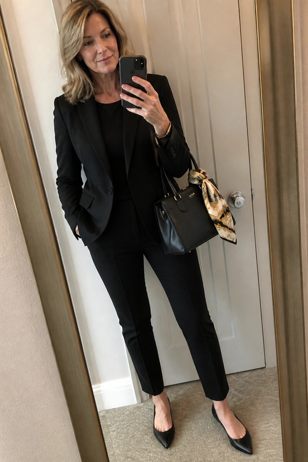 Woman in black pantsuit holding smartphone and leather handbag with patterned scarf