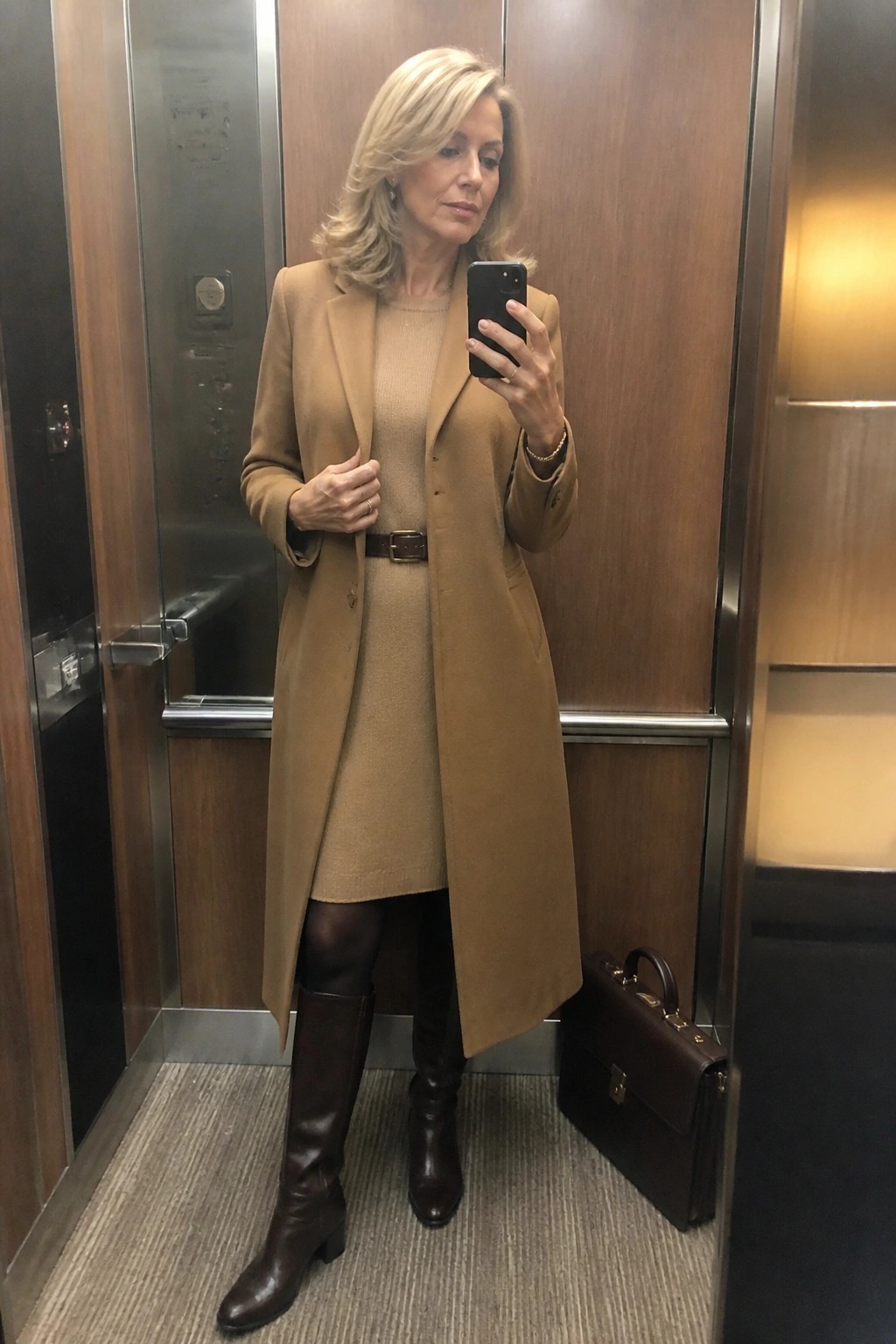 Woman in tan coat and brown boots taking selfie in elevator with briefcase