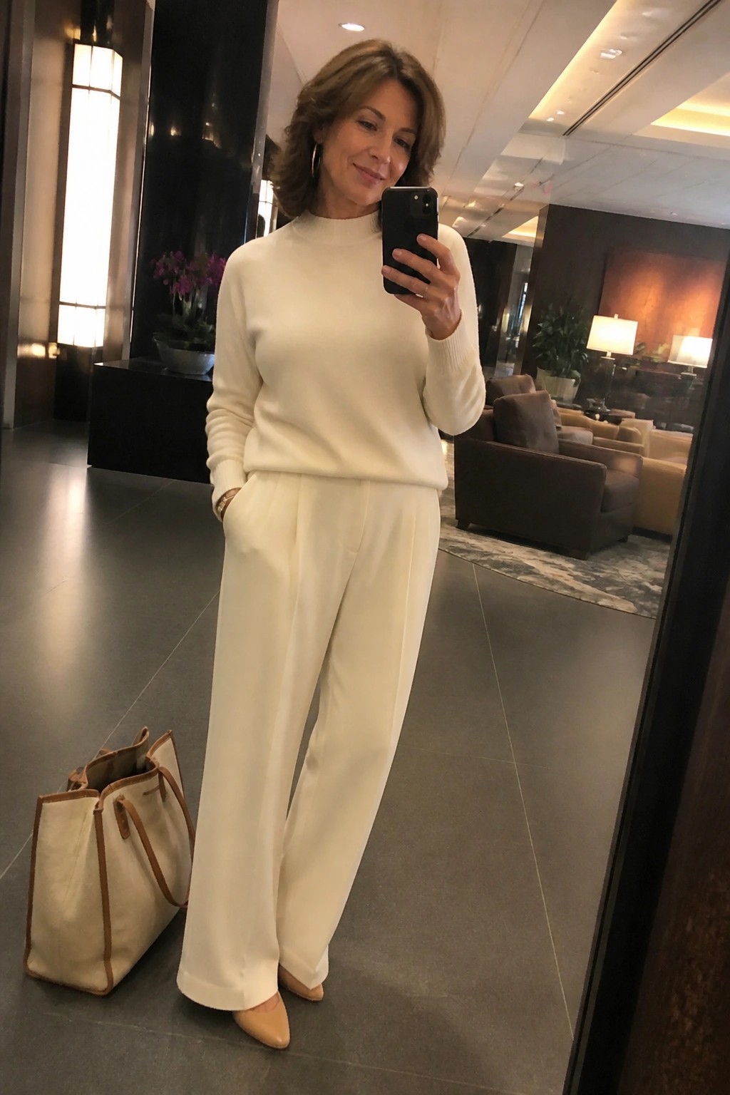 Woman in white outfit taking mirror selfie in lobby beside beige tote bag