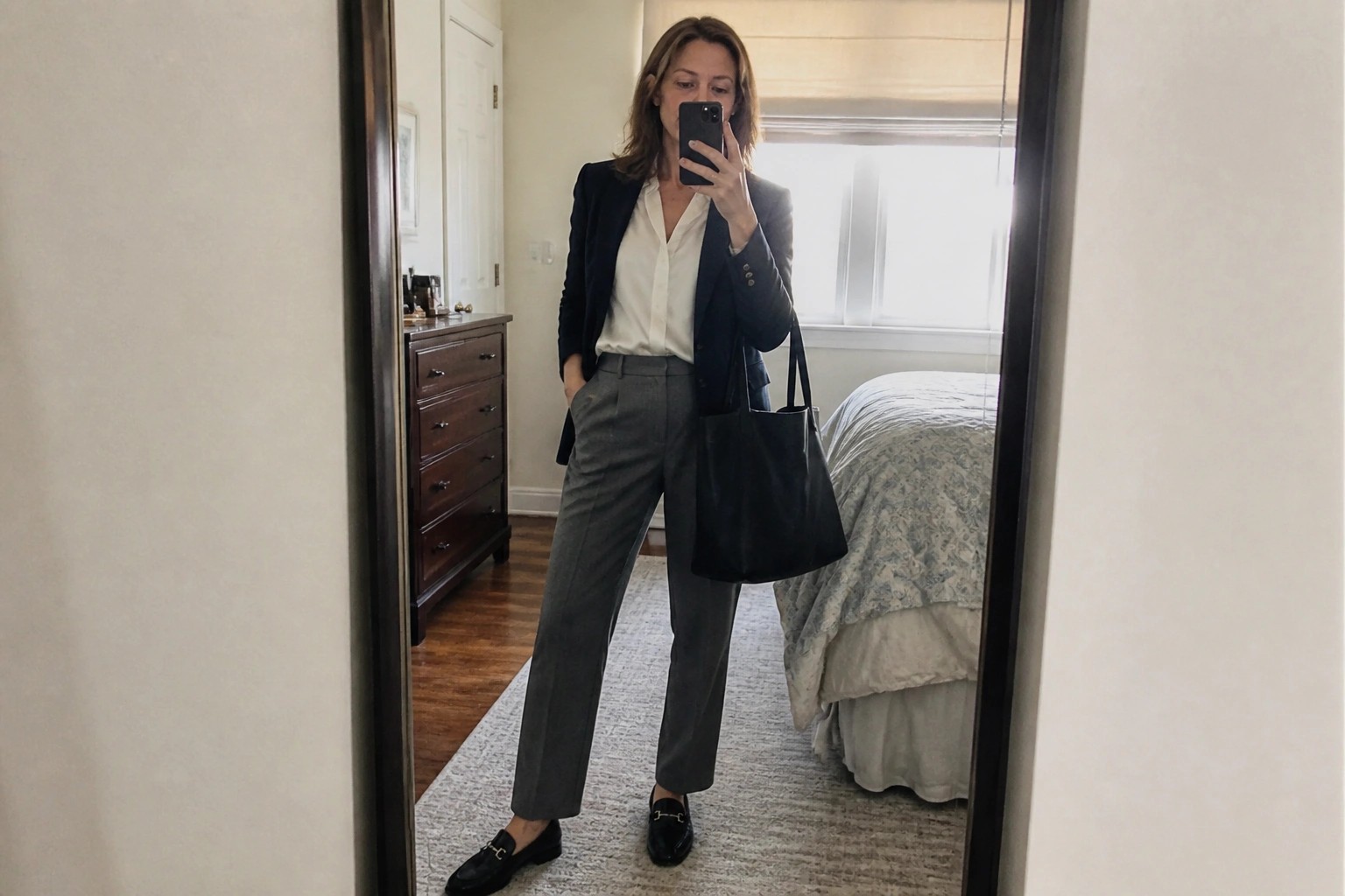 Woman in navy blazer and gray pants taking mirror selfie in bedroom