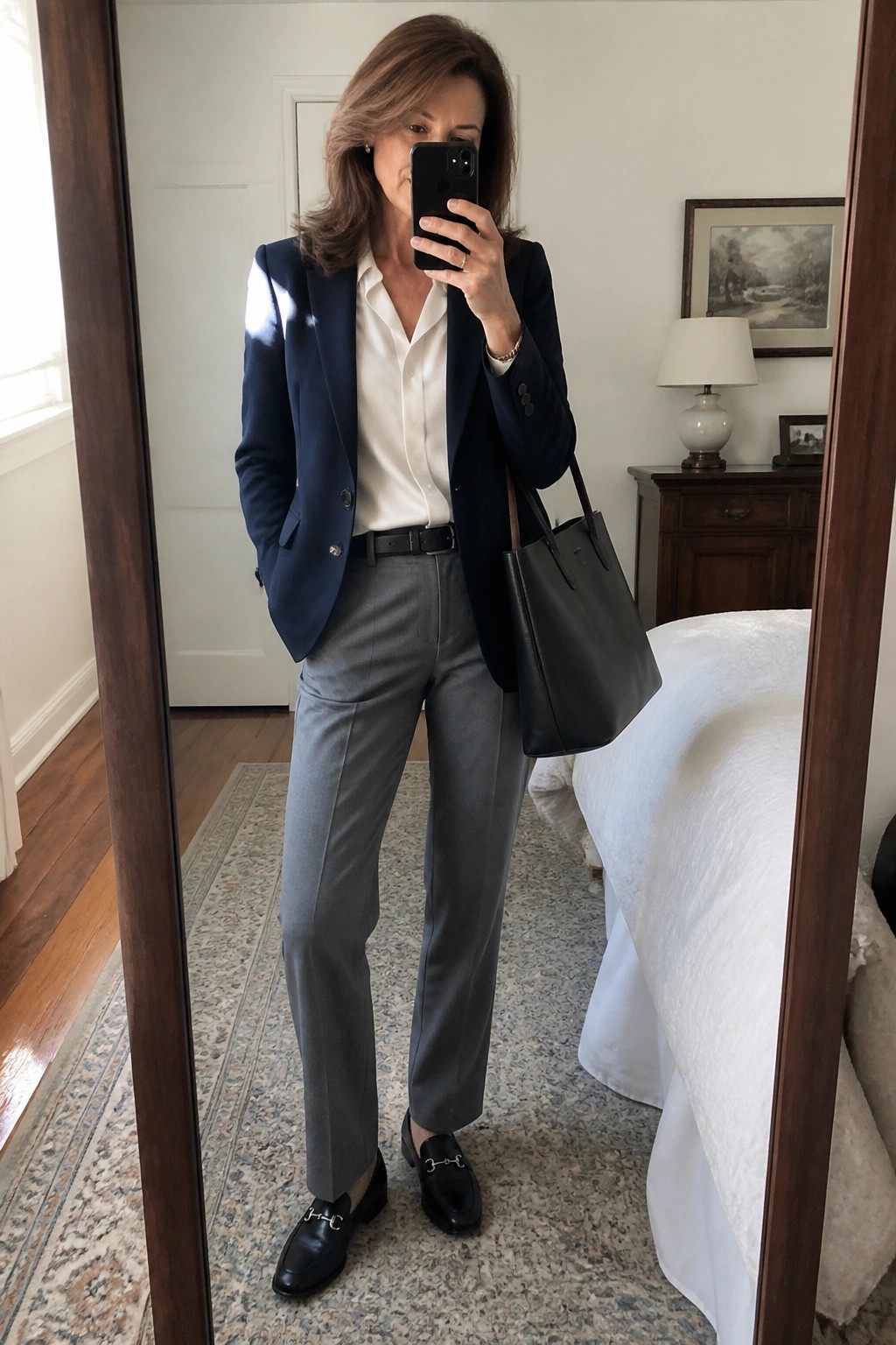 Woman in navy blazer and gray pants taking mirror selfie in bedroom