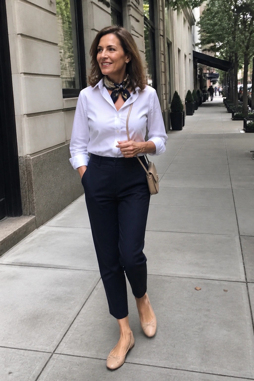 Woman over 50 in white button down shirt tucked into navy cropped trousers with patterned neck scarf beige crossbody bag and nude ballet flats standing on sidewalk with one hand in pocket.