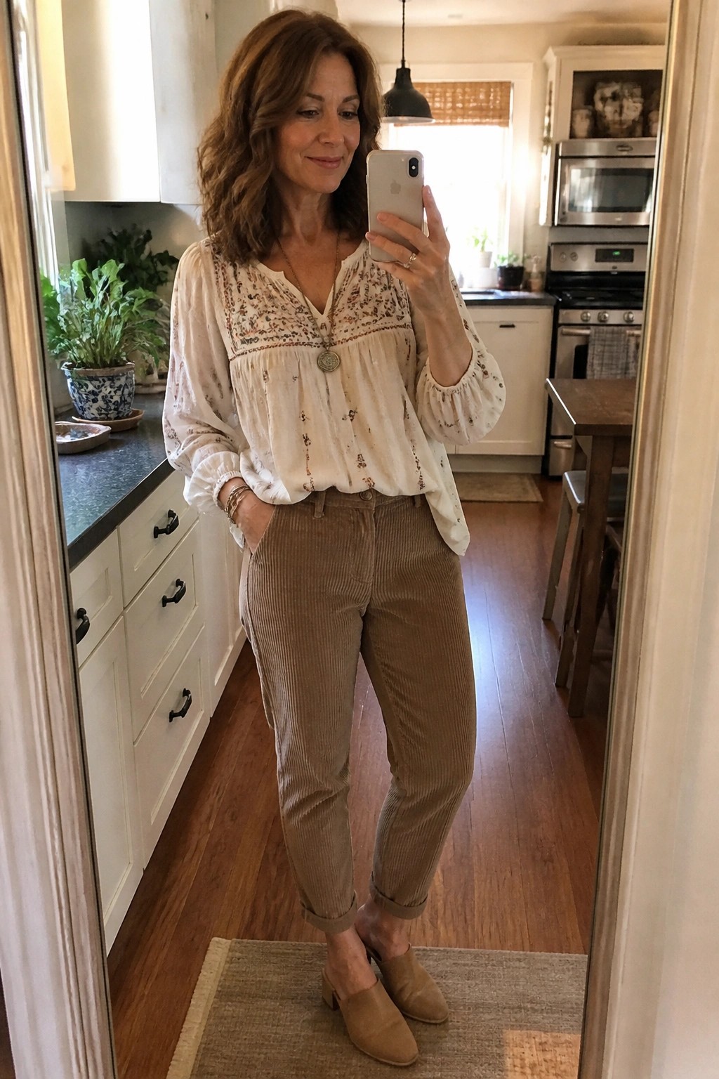 Woman over 50 in a cream floral embroidered blouse tucked into brown corduroy pants with beige mules and layered necklaces taking a mirror selfie in a kitchen setting
