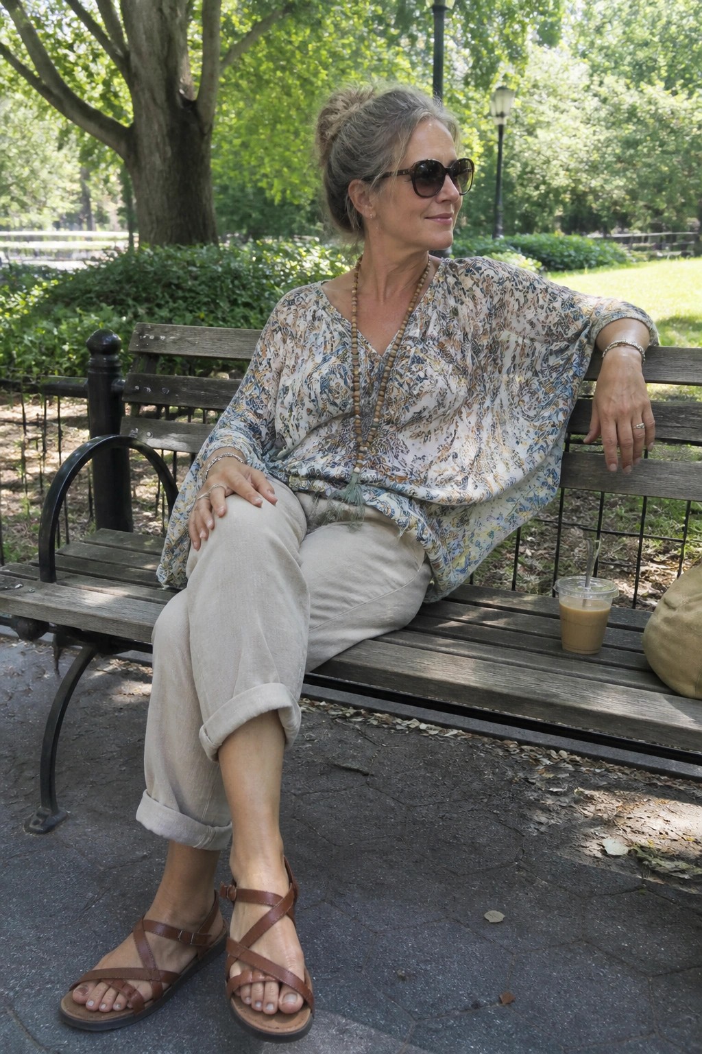 Woman in patterned blouse and linen pants sits on park bench with iced coffee