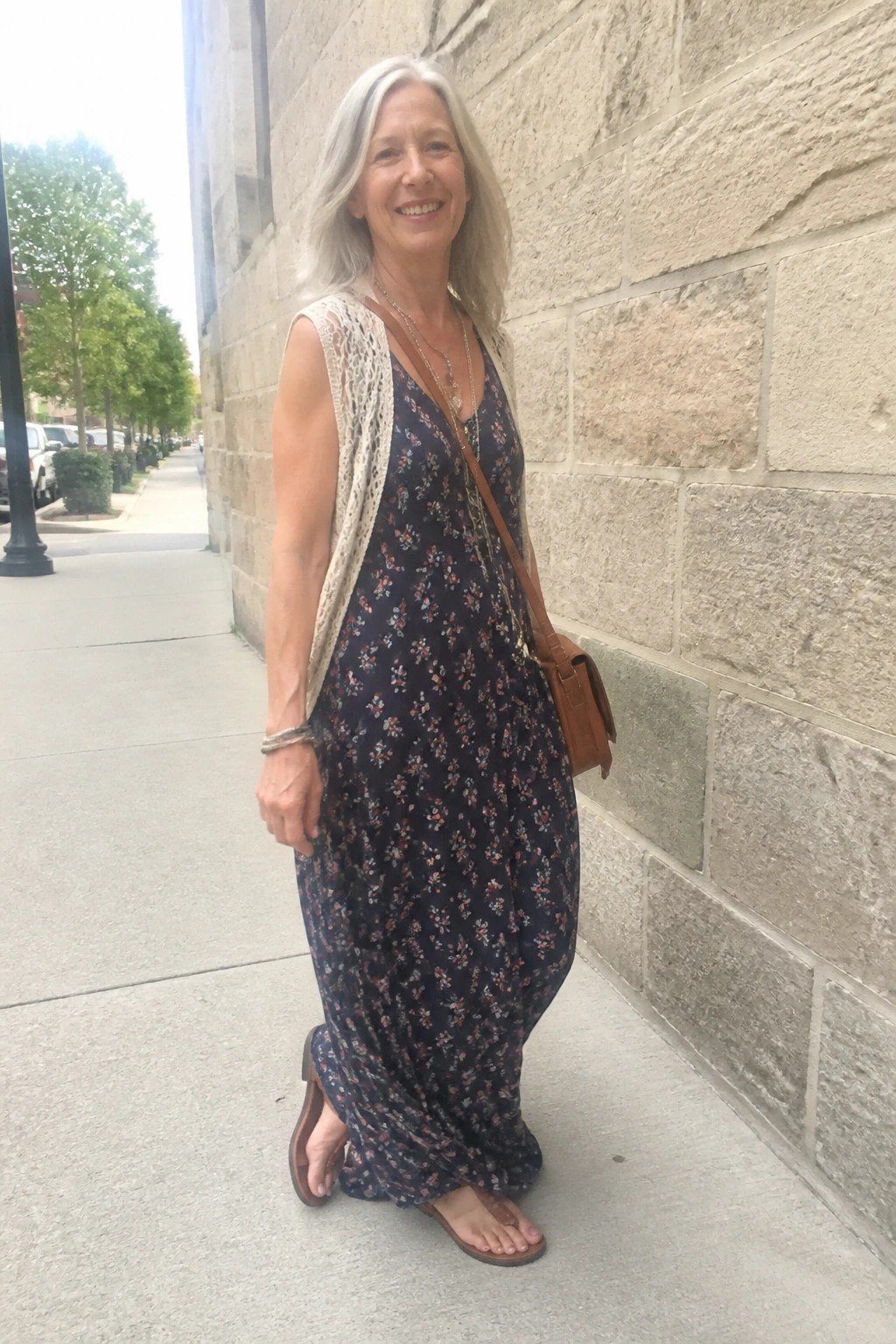 Woman with gray hair in navy floral dress and cream lace vest near stone wall